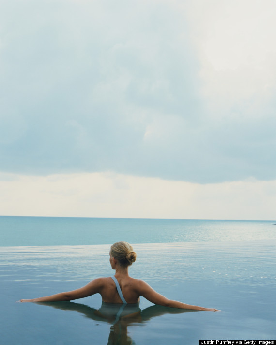 Young woman standing in pool by ocean, arms outstretched, rear view